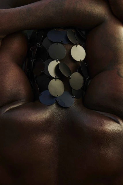 Upper body of a nude person with metallic brown skin. A beaded garment made of dark and light grey metal discs covers their chest.