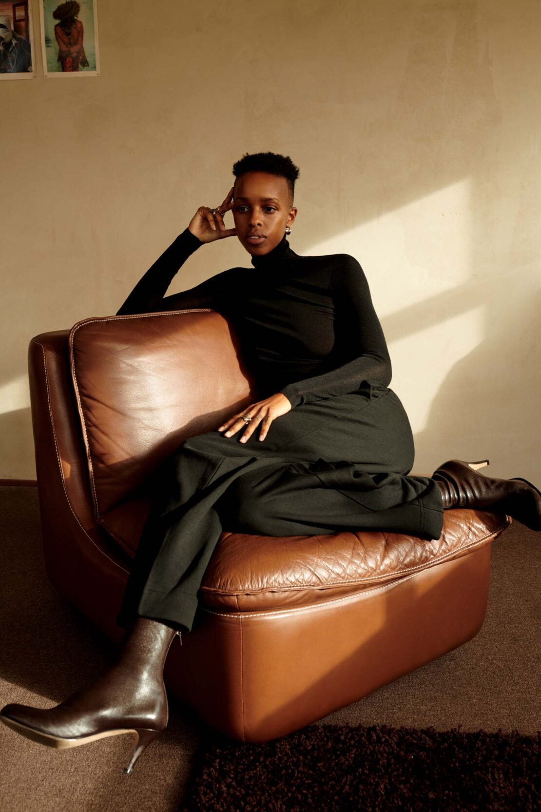 Stylish woman in black turtleneck and brown trousers, poses on vintage brown leather armchair, wearing heeled ankle boots indoors.