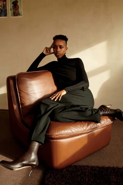 Stylish woman in black turtleneck and brown trousers, poses on vintage brown leather armchair, wearing heeled ankle boots indoors.