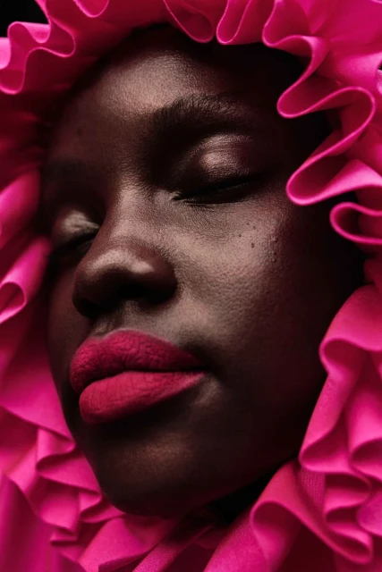 Close-up portrait of a dark-skinned woman with eyes closed, wearing pink lipstick and a ruffled pink collar in a studio setting.