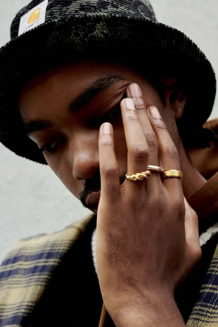 Close-up of a young adult Black man wearing rings and a bucket hat, gazing downward while partially covering his face with his hand.