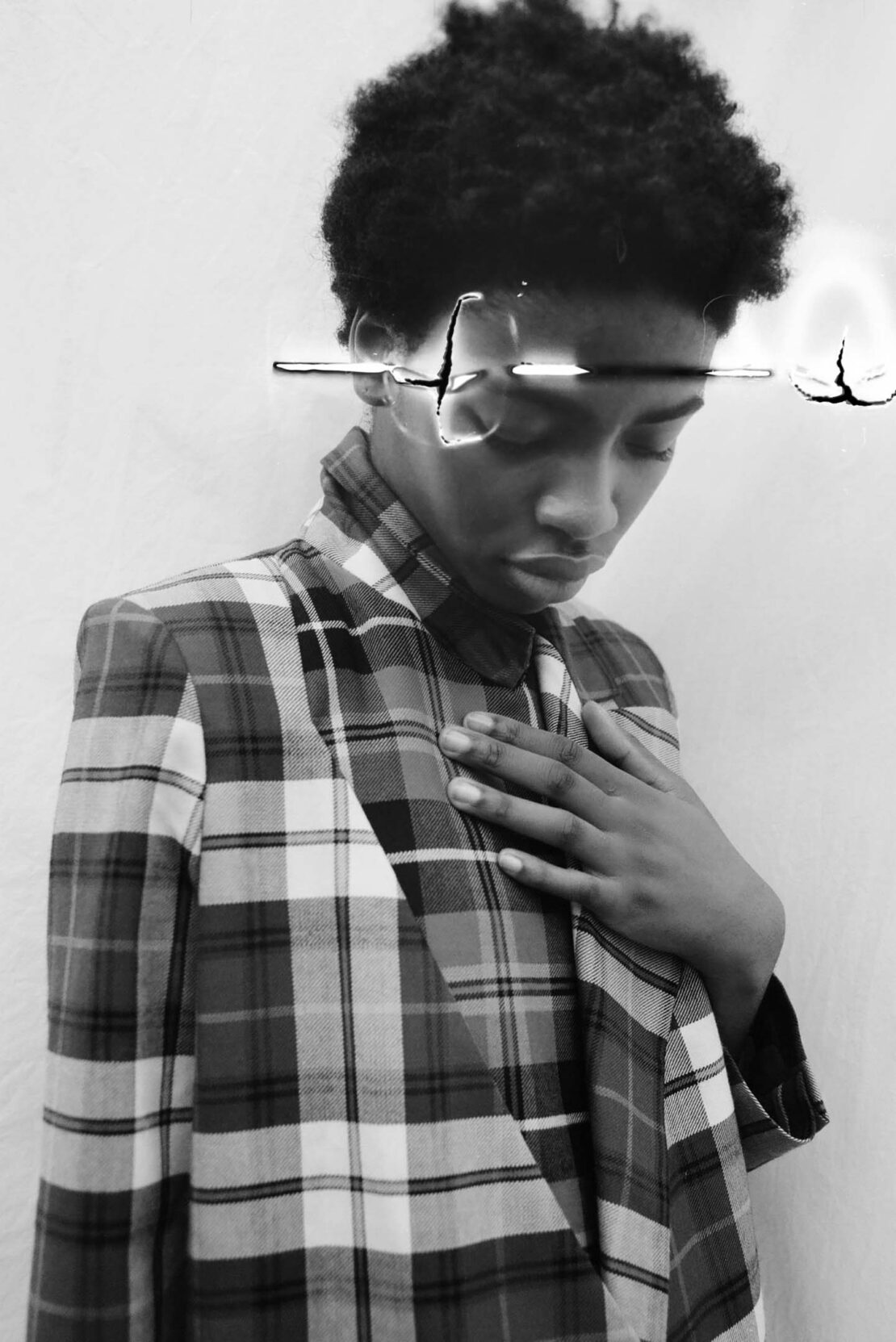 Monochrome portrait of an African-American person with an afro wearing a plaid blazer, hand on chest in an indoor environment.