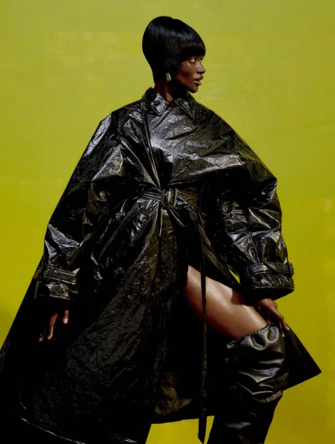Woman with short black hair poses in a black vinyl trench coat and black boots against a bright yellow-green backdrop in studio lighting.