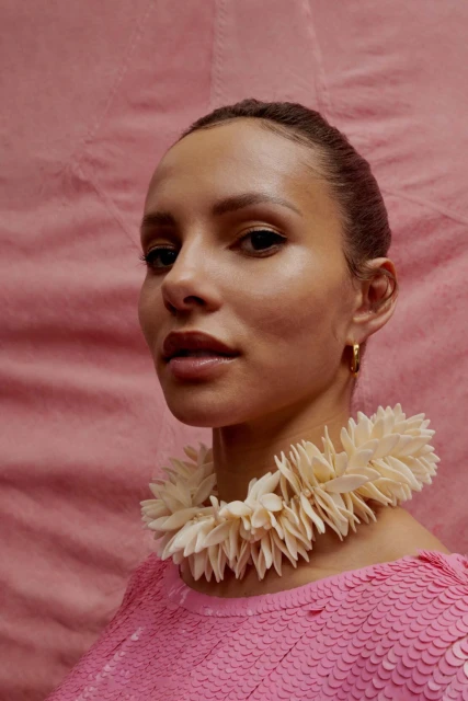 Portrait of a woman with light skin, wearing a pink sequined top and a white floral necklace against a textured pink background.