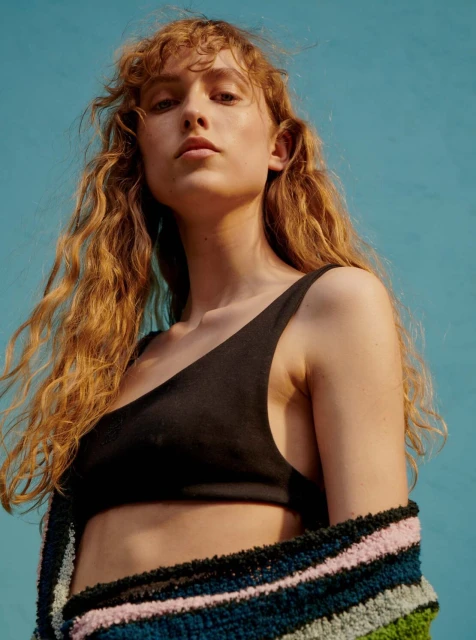Eye-level portrait of a young adult female model with auburn curly hair wearing a black top and colourful cardigan against a blue backdrop.