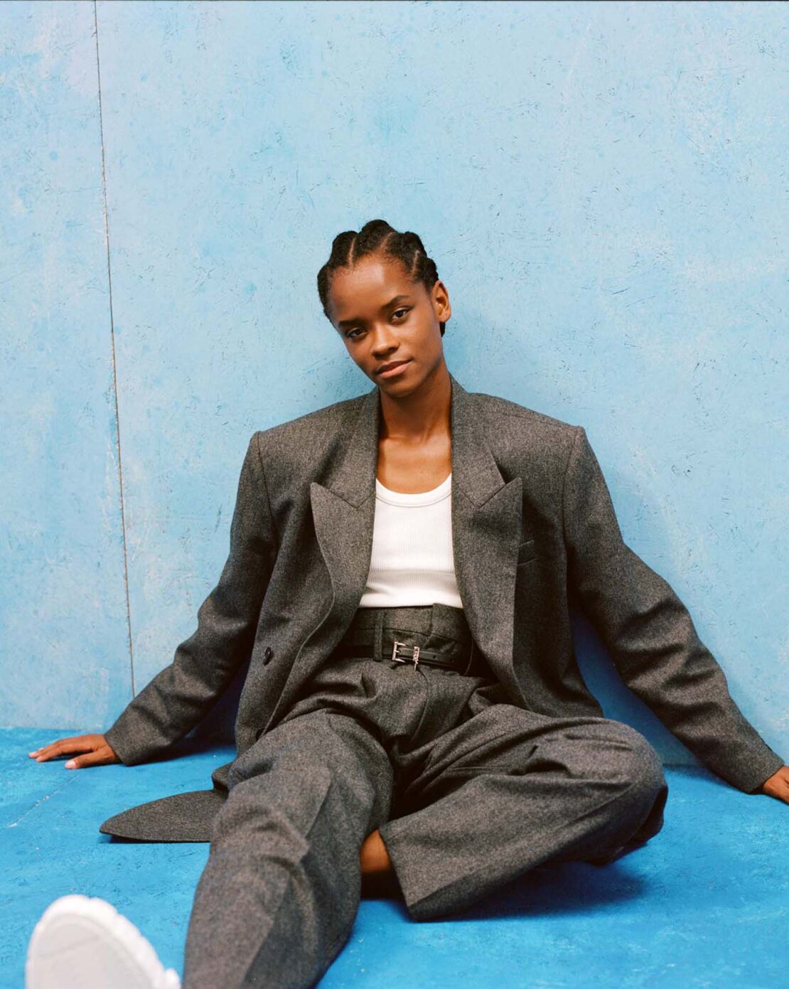 Portrait of a Black woman seated against a textured blue wall wearing a grey wool suit jacket and trousers