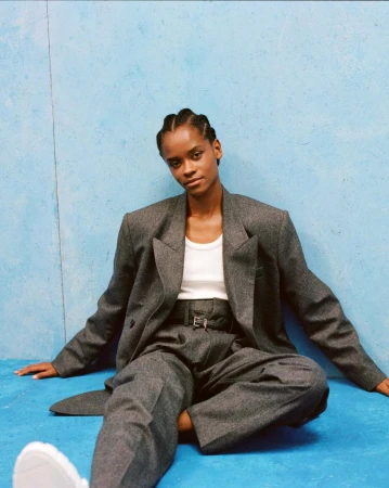 Portrait of a Black woman seated against a textured blue wall wearing a grey wool suit jacket and trousers