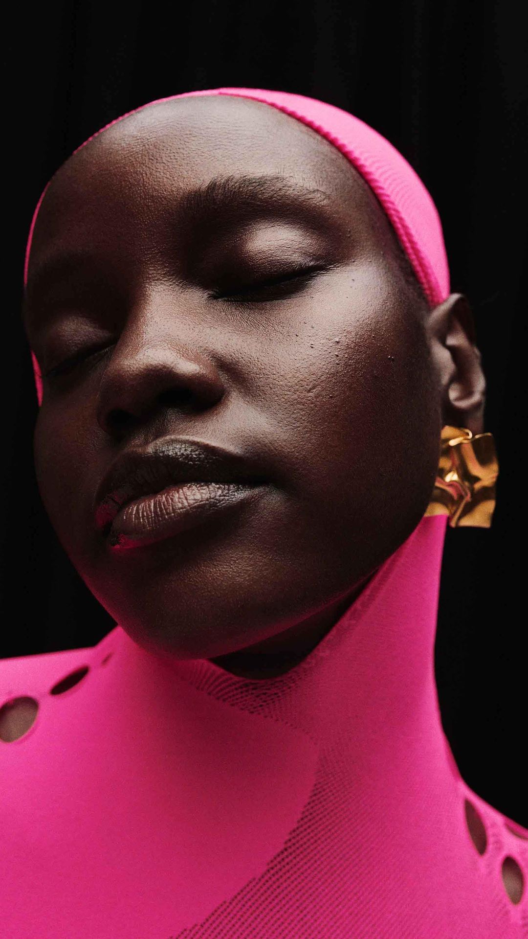 Close-up portrait of a woman with dark skin, eyes closed, wearing a pink headscarf, pink top, and gold earrings against a black background.