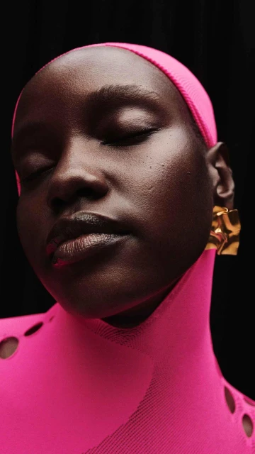 Close-up portrait of a woman with dark skin, eyes closed, wearing a pink headscarf, pink top, and gold earrings against a black background.