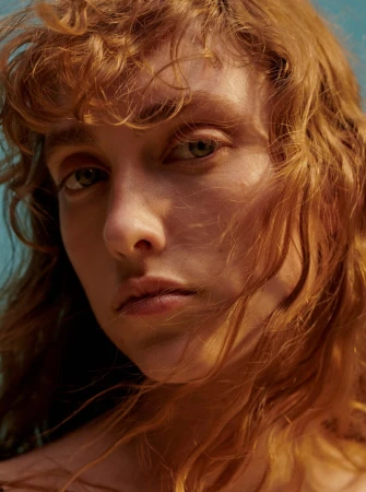 Close up portrait of a young woman with curly red hair and green eyes against a blue sky background
