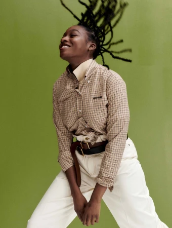 Black woman with dreadlocks flicking her head against a green background, wearing a plaid shirt and white trousers.