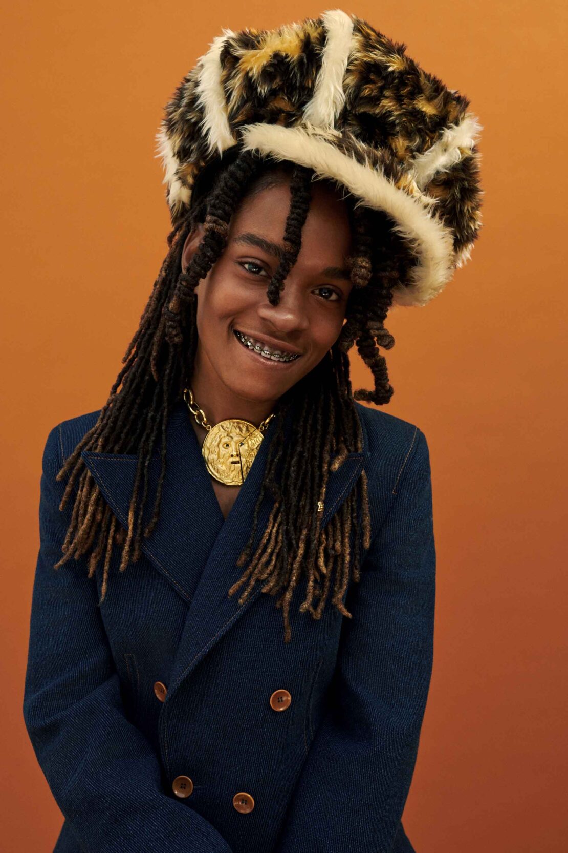 Portrait of a stylish adult with braces, wearing a leopard print hat, gold necklace, and blue denim jacket, posing against an orange background.