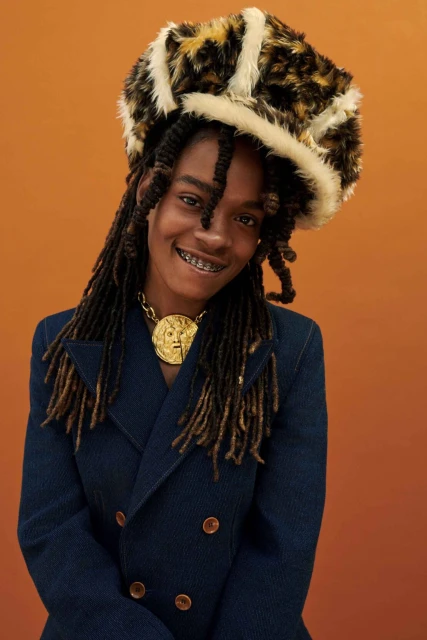 Portrait of a stylish adult with braces, wearing a leopard print hat, gold necklace, and blue denim jacket, posing against an orange background.