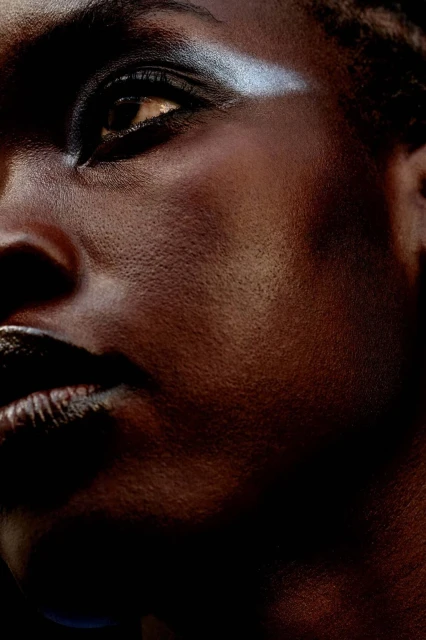 Close-up portrait of a Black woman with silver eyeshadow and dark lipstick, illuminated by dramatic lighting against a dark background.