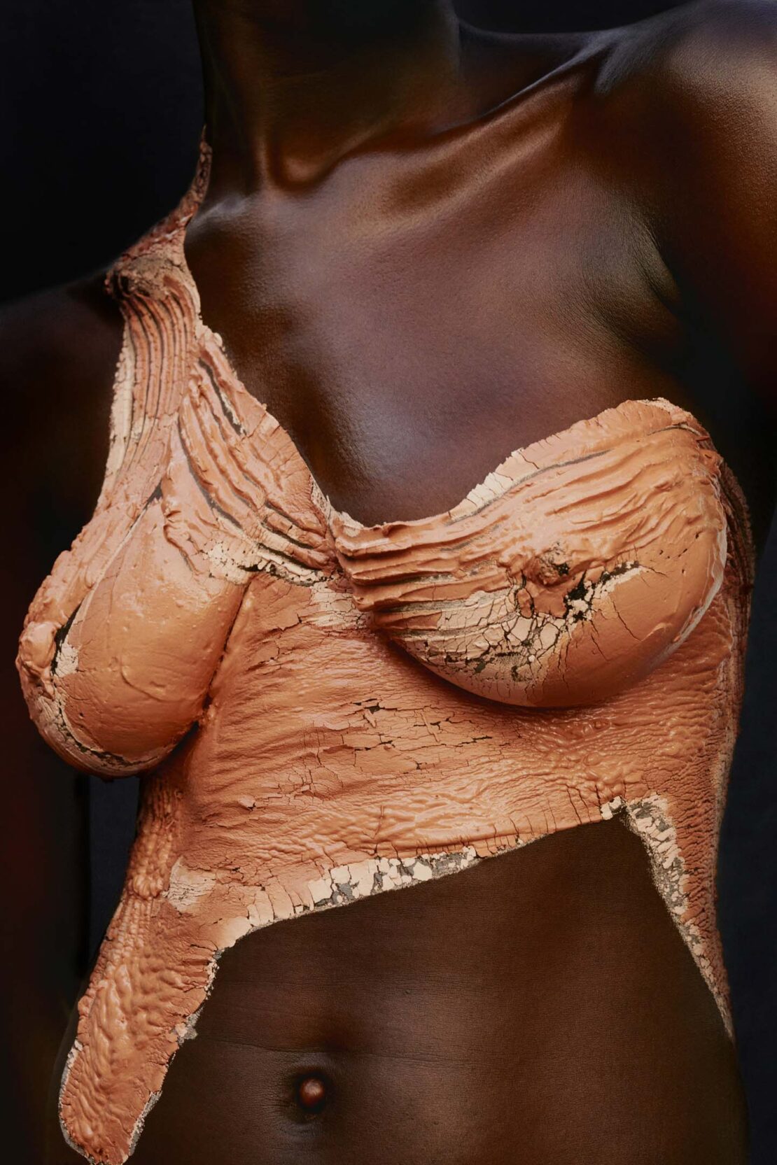 Close-up portrait of a dark-skinned adult female with a copper-colored clay-like material covering her chest and upper body against a dark backdrop.