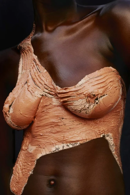 Close-up portrait of a dark-skinned adult female with a copper-colored clay-like material covering her chest and upper body against a dark backdrop.