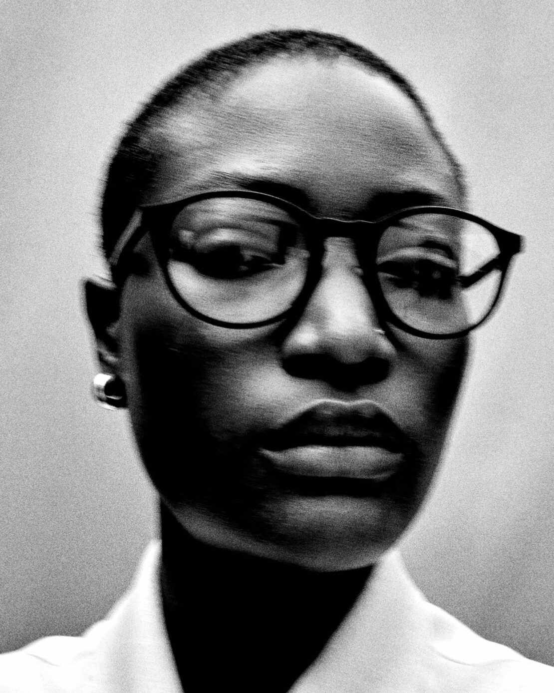Close-up black and white portrait of a bald adult woman wearing black glasses, silver earrings, and a white collared shirt against a plain background.