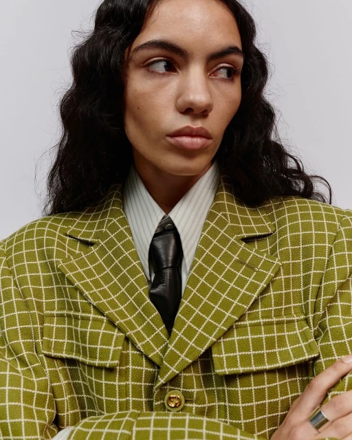 Woman in a patterned olive green blazer, wearing a dark tie and collared shirt, gazing off-camera with a neutral facial expression.