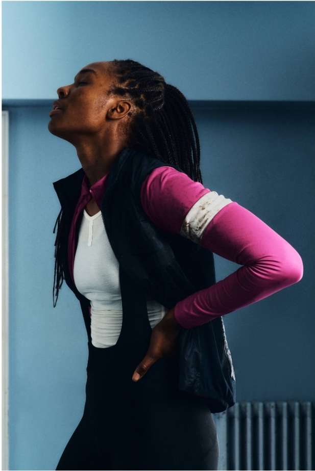 Profile of an athletic Black woman in activewear, looking up with hands on hips against a blue wall; fitness, health, sport, workout.