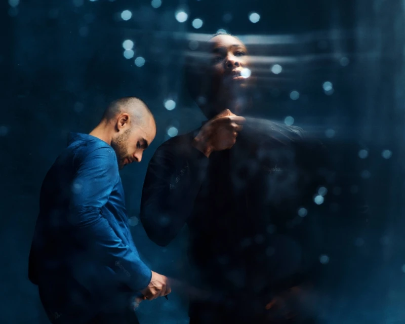 Two men in blue and black clothing stand in front of a dark background with bokeh, with one man's image appearing blurred.