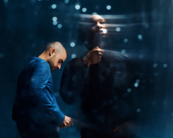 Two men in blue and black clothing stand in front of a dark background with bokeh, with one man's image appearing blurred.