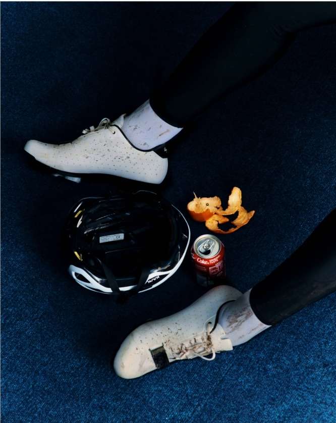 Close-up of cycling shoes, helmet, open soda can, and orange peel on a blue surface, suggesting a break during or after a bike ride.
