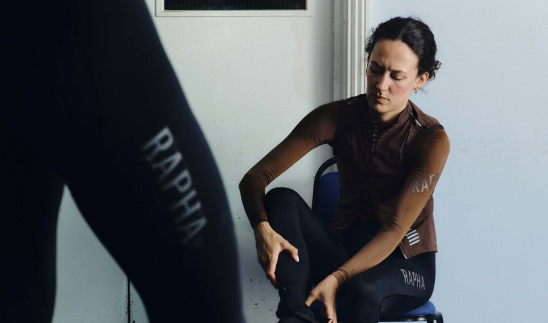 A cyclist in sports clothing sits on a chair indoors, with the Rapha logo visible on the clothing, in a brightly lit setting.