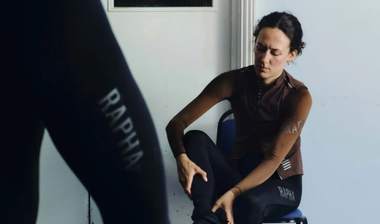 A cyclist in sports clothing sits on a chair indoors, with the Rapha logo visible on the clothing, in a brightly lit setting.