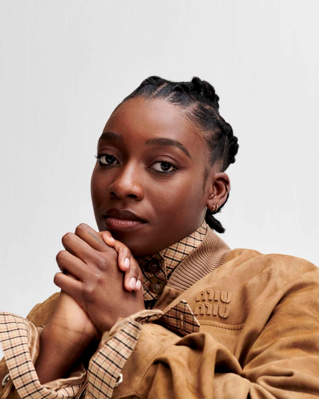 Close-up portrait of a woman with dark hair in braided style and wearing a brown jacket against a white background in a studio setting.