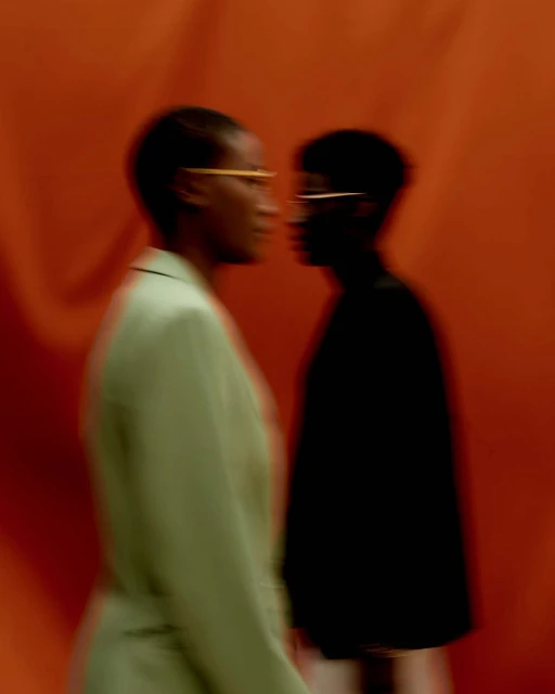 Two Black women in soft focus, face to face against a bold orange backdrop, wearing coats and glasses, creating an intimate fashion moment.