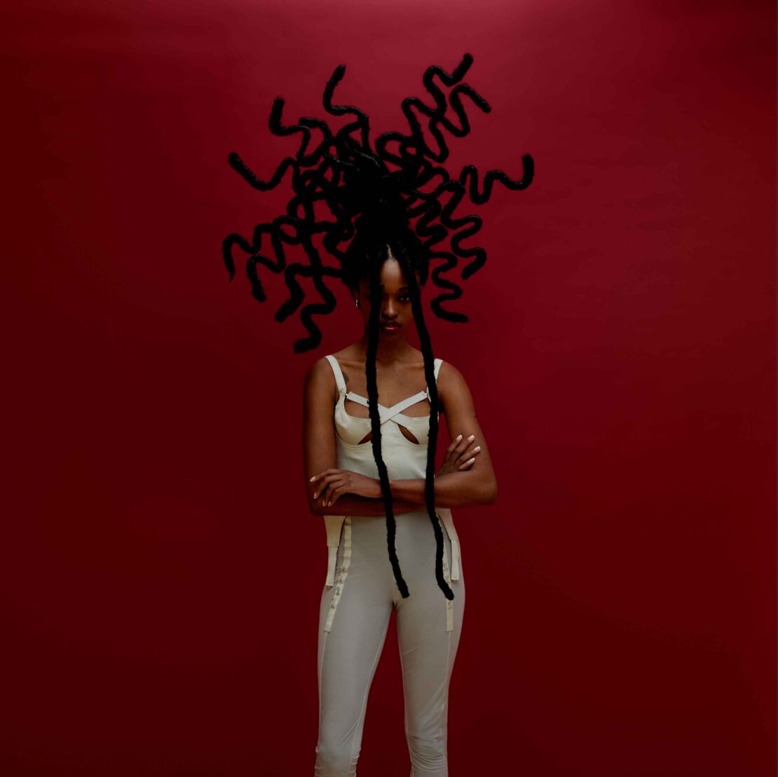 Portrait of a woman with intricately styled hair against a deep red background, posing in a light-colored top and pants.