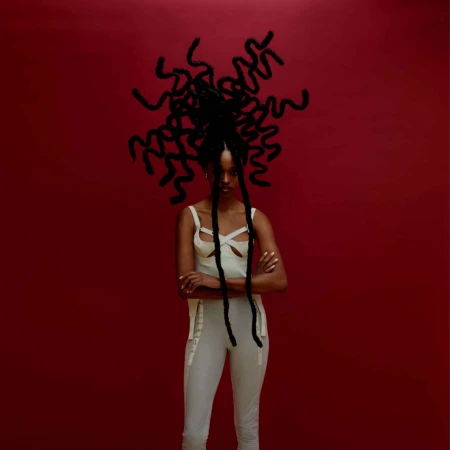 Portrait of a woman with intricately styled hair against a deep red background, posing in a light-colored top and pants.