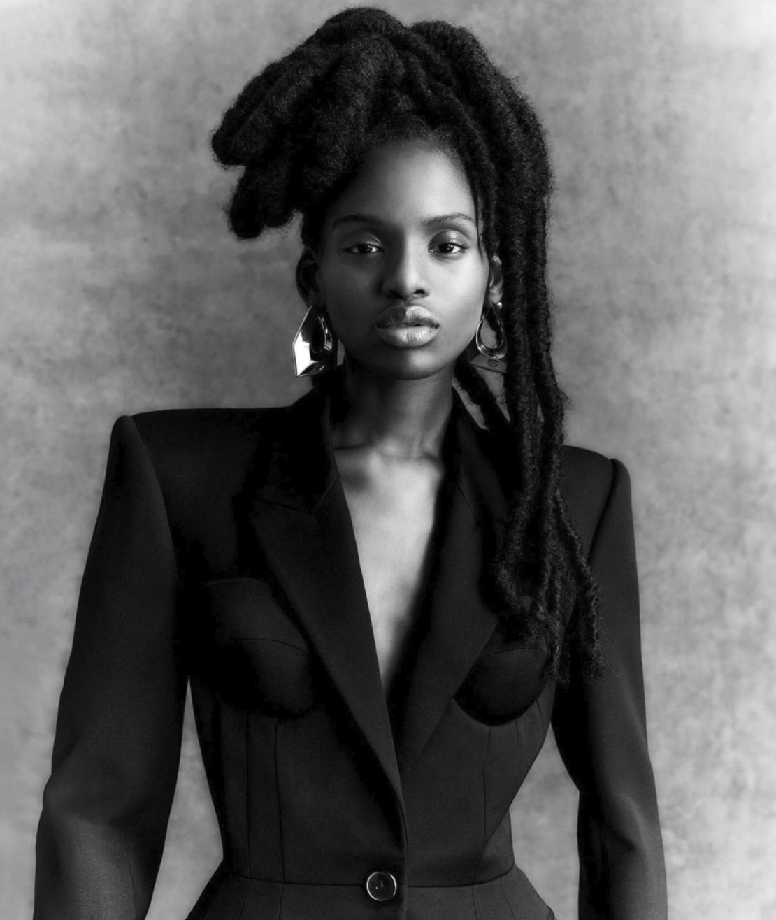 A striking black and white portrait showcases a Black woman with dreadlocks wearing a tailored blazer suit, set against a textured gray background.