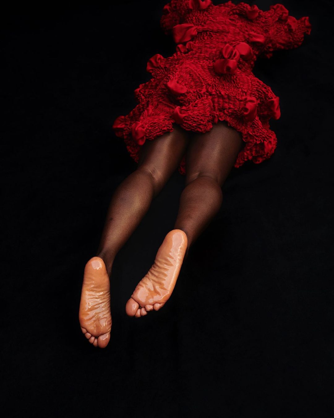An artistic studio shot features the lower torso and legs of a person with dark skin, clad in a textured red garment against a black backdrop.