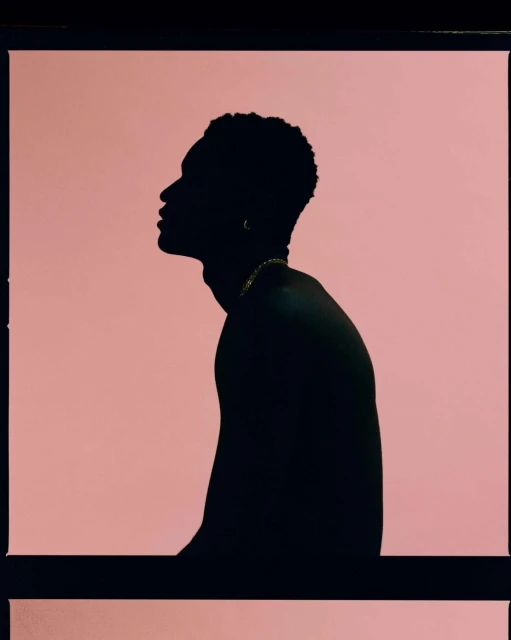 Silhouette of a bare-chested Black man against a pale pink background, framed by black border, highlighting his profile and gold necklace.