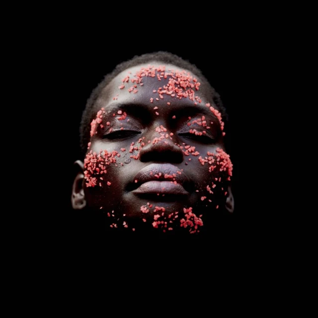 Close-up of a person with dark skin covered in small, red particles, eyes closed against a stark black background.