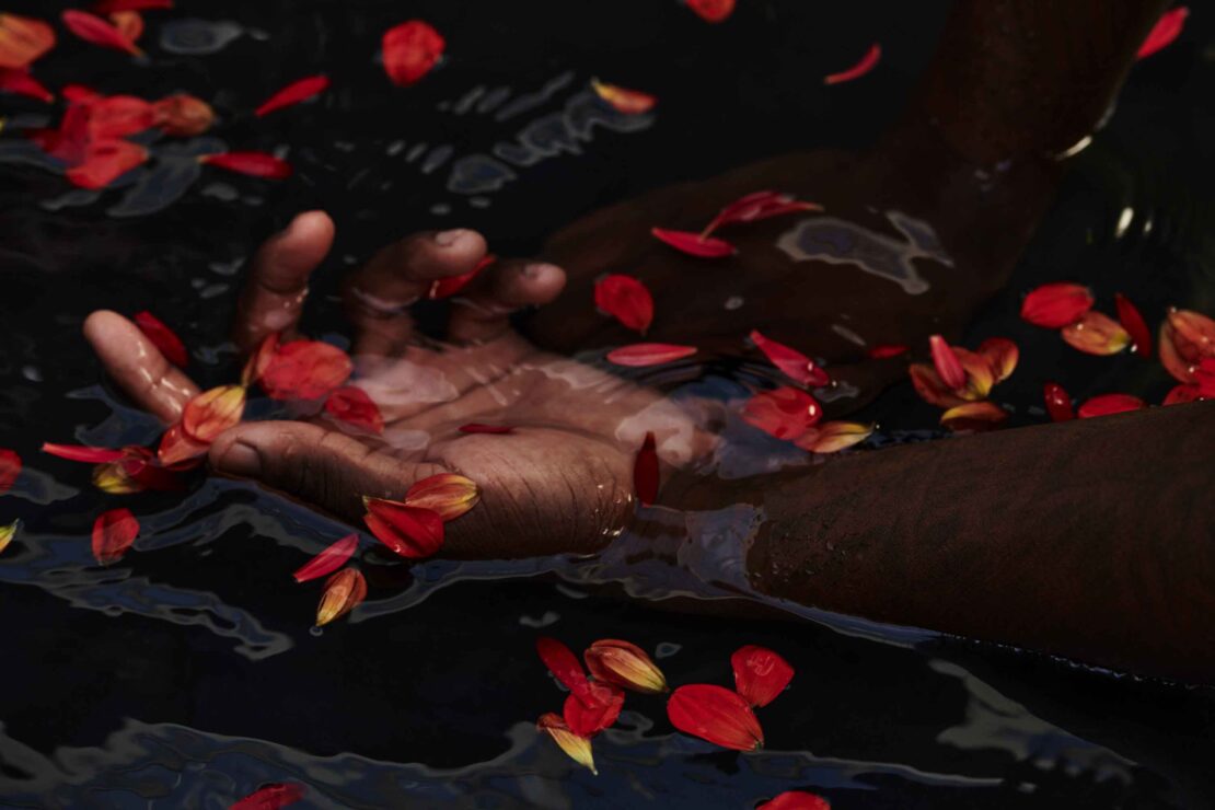 Close-up shot of adult hands immersed in dark water, surrounded by floating red and yellow flower petals creating a serene, moody image.