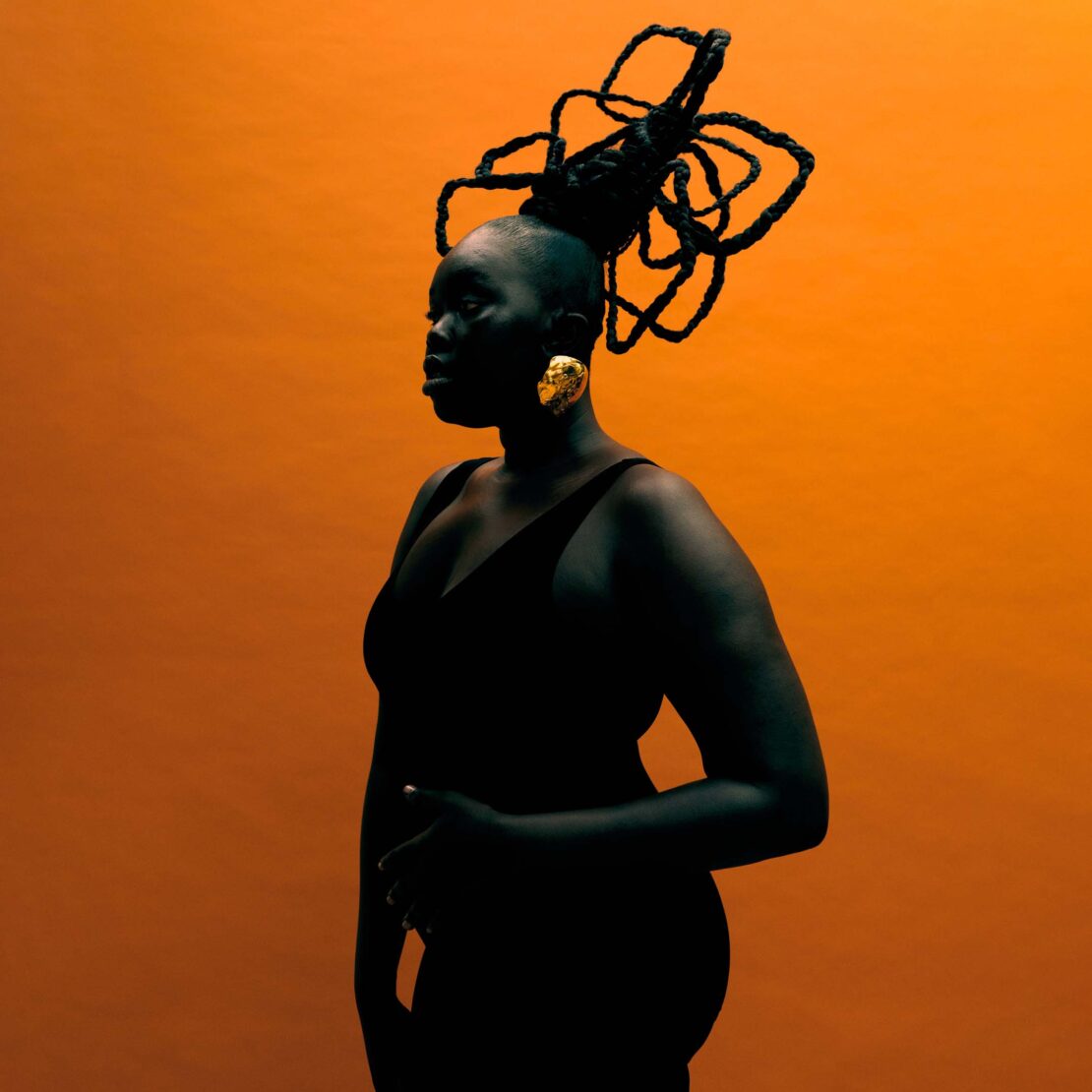 Striking portrait of a woman with a unique updo and gold earrings against a vibrant orange backdrop, showcasing beauty and style.
