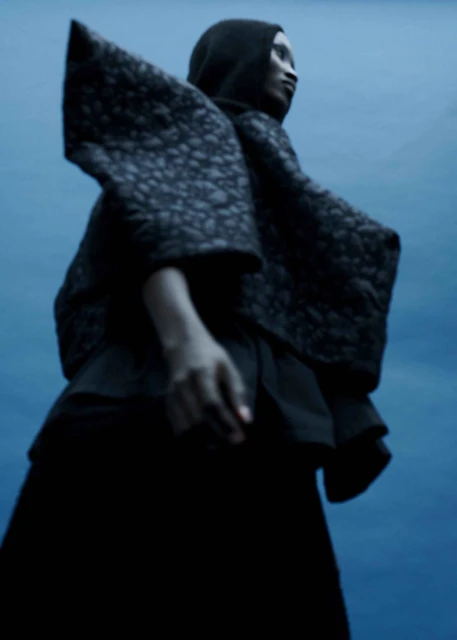 A dark-skinned adult female with a hooded shawl poses against a blue backdrop, in a low-angle medium shot for a fashion editorial.