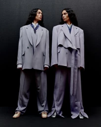 Two models with long dark hair wearing matching oversized lavender trouser suits and patterned neckties against a dark textured background