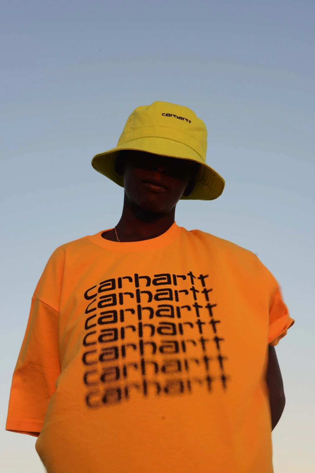 Low-angle shot of a person wearing a yellow bucket hat and orange graphic t-shirt against a clear sky.
