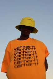 Low-angle shot of a person wearing a yellow bucket hat and orange graphic t-shirt against a clear sky.