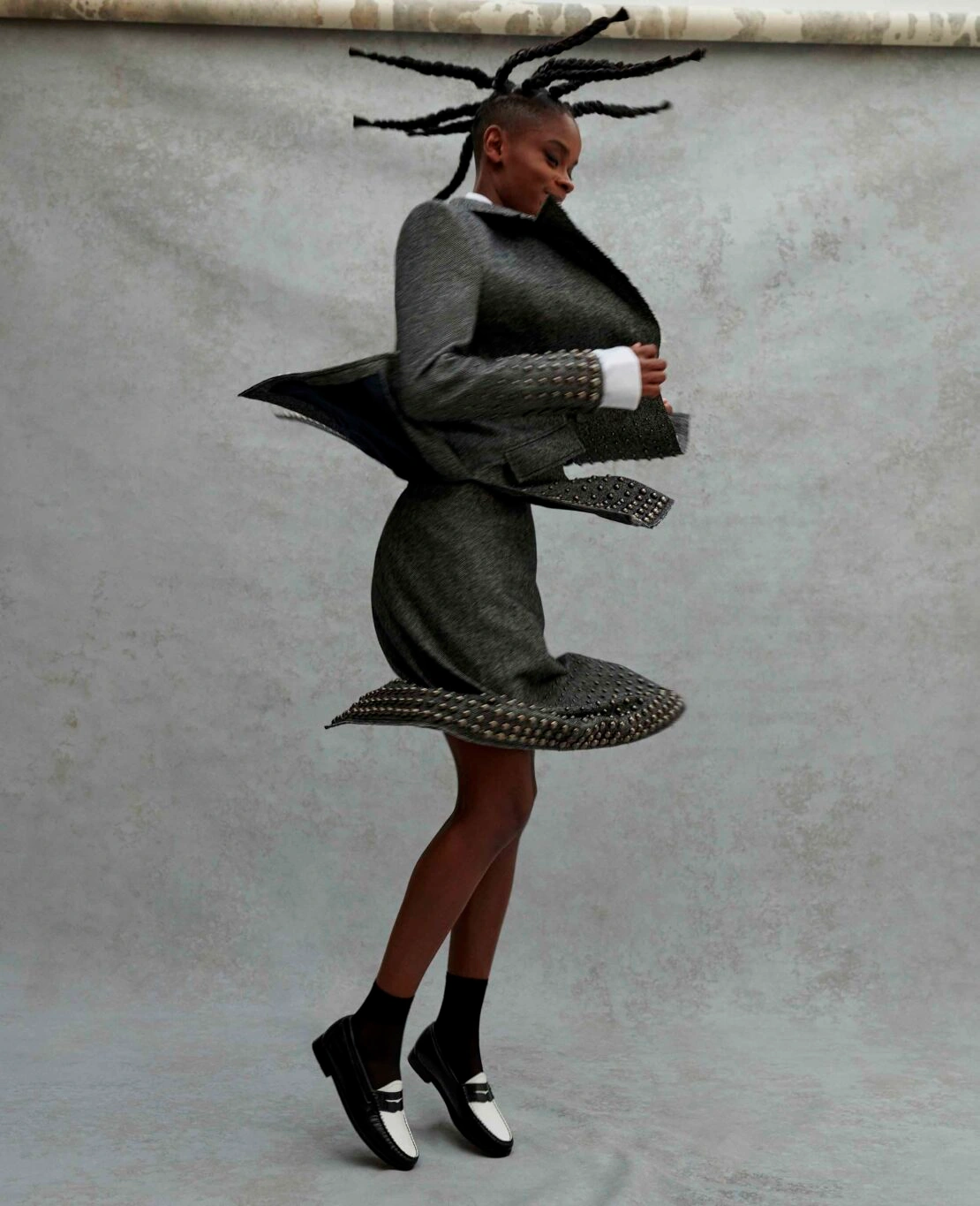 Black woman in motion wearing a gray textured skirt suit and two-tone loafers against a gray backdrop