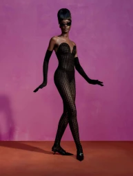 Black model in a textured black catsuit and matching gloves posing against a vibrant pink backdrop with a red floor.