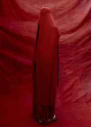 Figure completely draped in a long red hooded garment with fringed hem standing against a textured red background