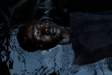 Close-up portrait of a Black woman floating in dark water with small rhinestones on her face and wearing a dark mesh top.