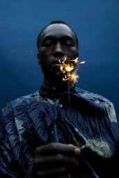 Black man holding a lit sparkler wearing a dark patterned shirt against a deep blue sky by Ekua King