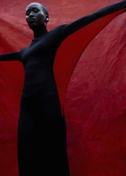 Low-angle shot of a black woman in a black turtleneck dress posing against a textured red background with outstretched arms.
