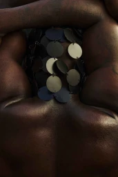 Close-up portrait of a dark-skinned person wearing a metallic disc necklace covering the face, photographed by Ekua King