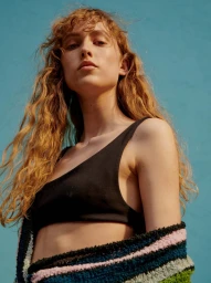 Low-angle portrait of a woman with curly blonde hair wearing a black top and striped cardigan against a blue sky background.